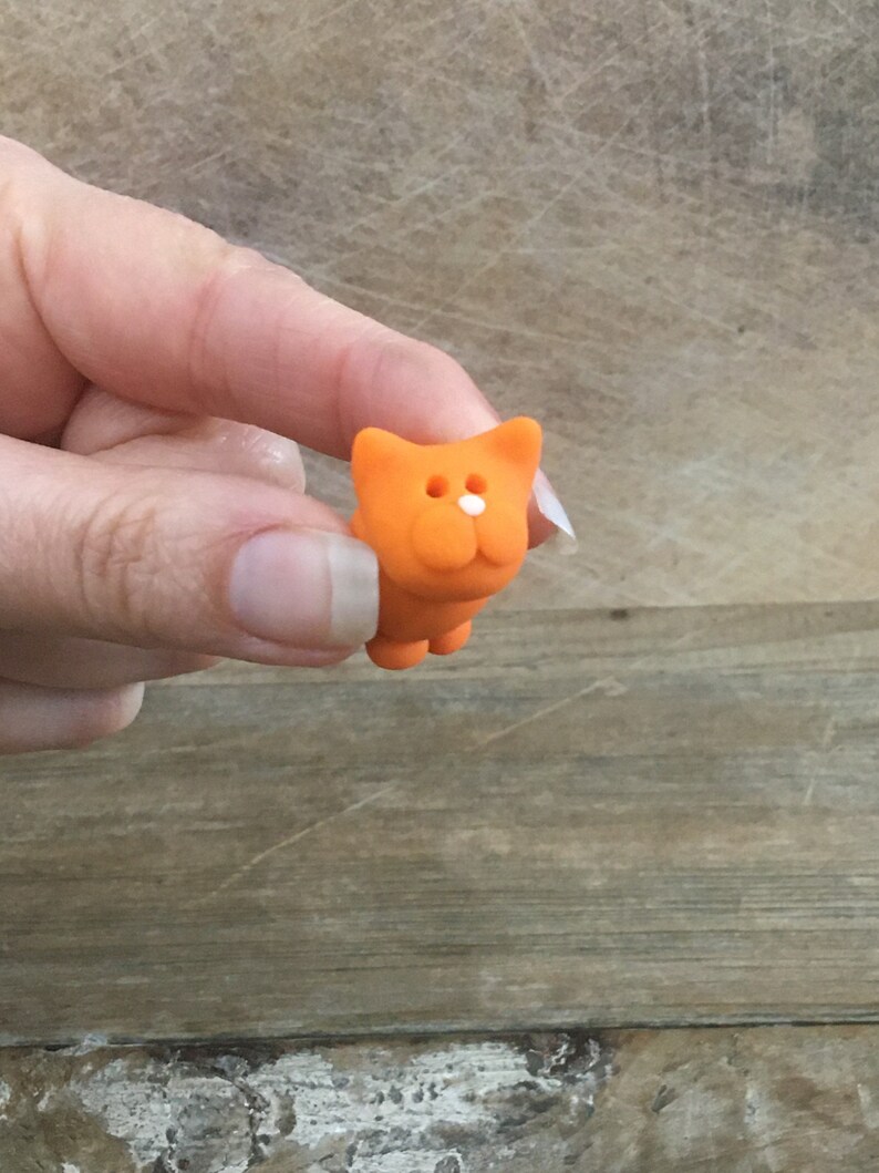 May include: A small, orange cat figurine with a white nose and two white eyes. The cat is held in a hand against a wooden background. The figurine has a simple, cartoonish design.