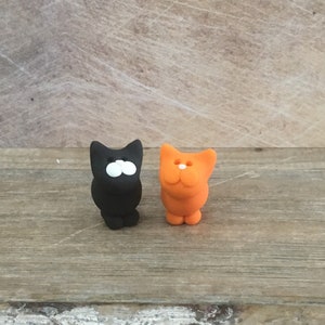 May include: Two small cat figurines. One is black with white eyes, the other is orange. Both have a simple, cartoonish design. They are standing on a wooden surface.