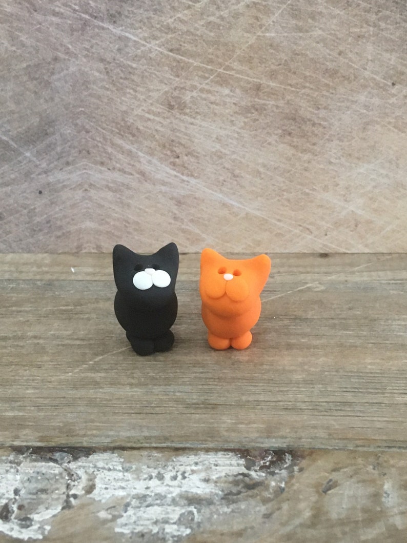 May include: Two small cat figurines. One is black with white eyes, the other is orange. Both have a simple, cartoonish design. They are standing on a wooden surface.