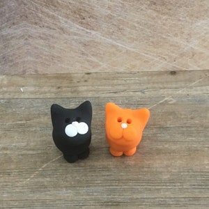 May include: Two small cat figurines. One is black with white facial features, the other is orange with white facial features. Both are standing upright on a wooden surface. The figurines have a simple, cartoonish design.