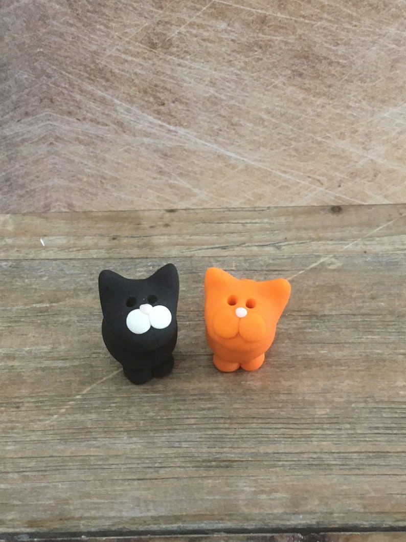 May include: Two small cat figurines. One is black with white facial features, the other is orange with white facial features. Both are standing upright on a wooden surface. The figurines have a simple, cartoonish design.