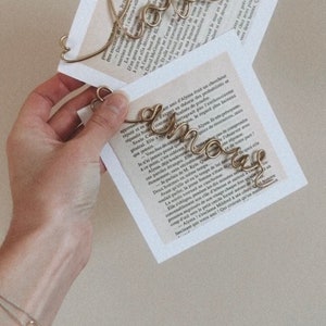 Op de afbeelding: Een gouden draadbeeld van het woord "Love" wordt in een hand gehouden. Het beeld is geplaatst op een boek met tekst in het Frans. Het woord "Love" is in een cursief schrift.