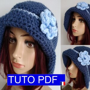 May include: A blue crocheted hat with a light blue crocheted flower. The hat has a wide brim and a scalloped edge. The hat is shown on a mannequin with red hair. The image also includes the text "TUTO PDF".