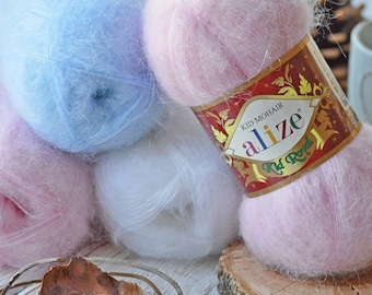 May include: A selection of Alize Kid Mohair yarn skeins in pastel shades of blue, pink, and white. One pink skein is displayed on a wooden slice. The yarn is labelled 'Alize'. The fluffy texture suggests it's suitable for knitting or crochet.