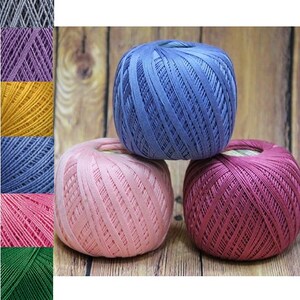 May include: Three balls of colorful crochet thread in shades of blue, pink, and burgundy are stacked on a wooden surface. The thread is tightly wound, and the colors are vibrant. A color chart of various thread colors is on the left.