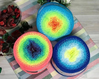 May include: Three balls of colourful yarn in shades of blue, green, yellow, and pink are displayed on a striped fabric. The yarn has a gradient pattern, creating a vibrant display. A bouquet of white and purple flowers is in the background.