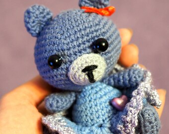 May include: A handmade crochet teddy bear in shades of blue, with a small red bow and black eyes. The bear is wearing a blue dress with a heart-shaped embellishment. The toy is held in a hand.