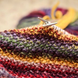May include: Close-up of knitting needles in a colourful, textured knitted project. The yarn is a mix of red, orange, yellow, green, and purple. A ball of yarn in similar colours is in the background.