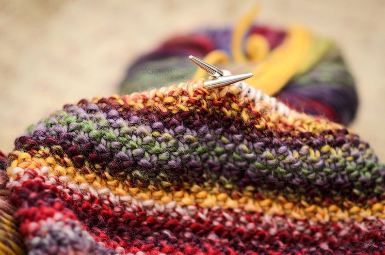 May include: Close-up of knitting needles in a colourful, textured knitted project. The yarn is a mix of red, orange, yellow, green, and purple. A ball of yarn in similar colours is in the background.