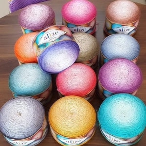 Puede incluir: Una colección de ovillos de hilo Alize Diva en varios colores, incluyendo rosa, morado, naranja y turquesa. Cada ovillo está cuidadosamente empaquetado, mostrando el efecto de color degradado del hilo. El hilo se exhibe sobre una superficie de madera.