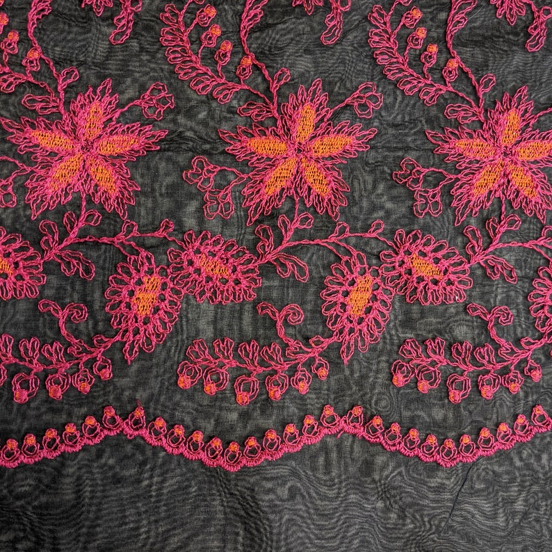 May include: Close-up of a fabric with a floral embroidered design. The embroidery features bright pink flowers with orange centers and stems, set against a dark gray, textured background. The bottom edge has a scalloped border.
