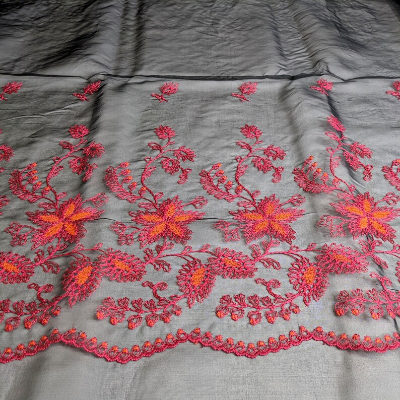 May include: Black sheer fabric with a floral embroidered border. The embroidery features vibrant pink and orange flowers and foliage. The fabric has a subtle sheen, and the floral design is detailed and intricate, suitable for apparel or decorative projects.