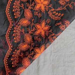 May include: Black fabric with intricate orange floral embroidery. The design features detailed flowers, leaves, and vines, creating a rich, textured pattern. The embroidery is highlighted against the dark background, showcasing the craftsmanship.