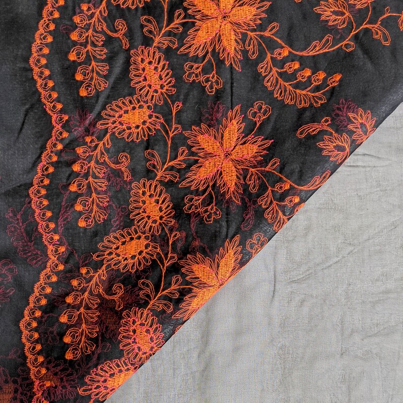 May include: Black fabric with intricate orange floral embroidery. The design features detailed flowers, leaves, and vines, creating a rich, textured pattern. The embroidery is highlighted against the dark background, showcasing the craftsmanship.