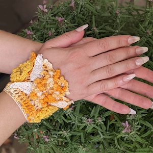 May include: A crocheted bracelet with a floral design in shades of yellow and white. The bracelet is worn on a person's wrist.