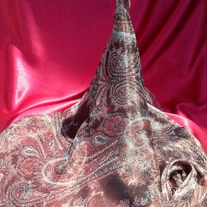 Könnte beinhalten: Ein brauner Schal mit Paisley-Muster und roten und weißen Akzenten liegt auf einer pinkfarbenen Satin-Oberfläche. Der Schal ist gefaltet und verdreht, wodurch eine kegelförmige Form entsteht. Der Stoff hat eine glänzende, seidige Textur.