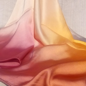 May include: A silk scarf in a gradient of colours, shifting from white to peach, then to pink, and finally to orange. The fabric is draped, highlighting its smooth texture and colour variations. The scarf appears to be made of a lightweight material.