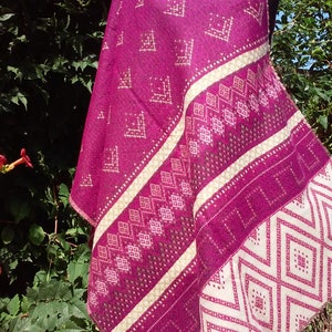 Può includere: Uno scialle magenta con motivi geometrici e strisce color crema. Lo scialle è drappeggiato su un manichino, mostrando il suo design intricato. I bordi sono rifiniti con frange. Lo sfondo presenta un rigoglioso fogliame verde.