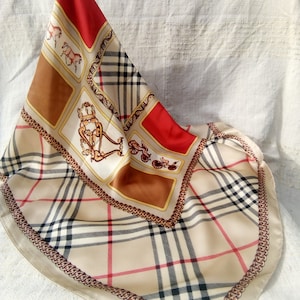 May include: A silk scarf in tan, red, and brown. It has a plaid design, horse and carriage illustrations, and a chain border. The scarf is folded, displaying its pattern and details. The measurements are in centimeters.
