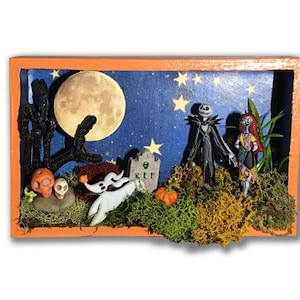 May include: A 3D diorama featuring Jack Skellington and Sally from The Nightmare Before Christmas. The diorama is set against a blue night sky with a full moon and stars. There is a small tombstone that reads "RIP".
