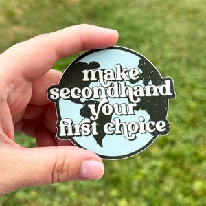 May include: A round sticker with a light blue background and a black outline of the Earth. The sticker has the words "make secondhand your first choice" in white, bold font.