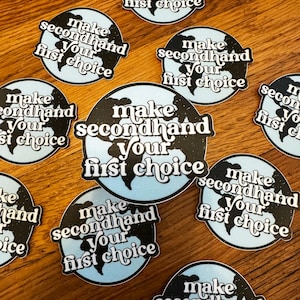 May include: A collection of circular stickers with a light blue globe design and the text "make secondhand your first choice" in a white, retro font. The stickers are scattered on a wooden surface.
