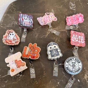 May include: A collection of eight Halloween-themed badge reels featuring various designs including ghosts, skeletons, and medical-themed text such as CNA, LAB, CODE BOO, and DIDN'T STAB ANYONE TODAY. Each reel has a retractable cord and a clear plastic clip.
