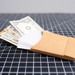 May include: A brown cardholder with a stack of white cards and several US dollar bills fanned out. The cardholder is made of cardboard and is open, revealing the cards. The background is a black grid.