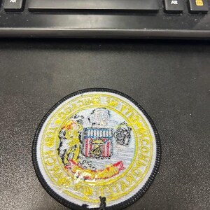 May include: A round, white and yellow embroidered patch with a black border. The patch features a seal with an eagle, a shield, and the words "United States Postal Service".