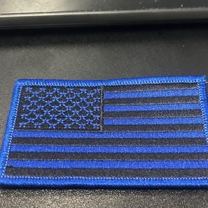 May include: A blue and black embroidered American flag patch. The stars are black and the stripes are blue.