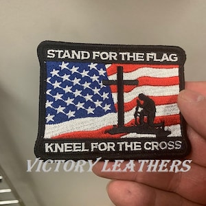 May include: Embroidered patch with an American flag and a cross. The text reads "Stand for the flag, kneel for the cross". The patch is black with white and red stitching. The patch is made by Victory Leathers.