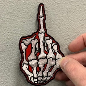 May include: A red and white embroidered patch of a skeleton hand giving the middle finger.