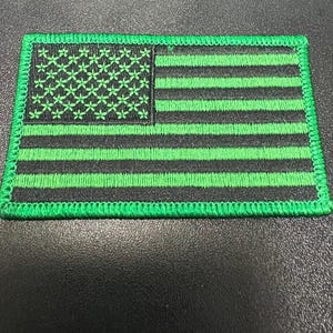 Puede incluir: Parche bordado con un diseño estilizado de la bandera estadounidense. La bandera tiene rayas negras y verdes y un campo de patrones florales verdes. El parche es rectangular con un borde verde.