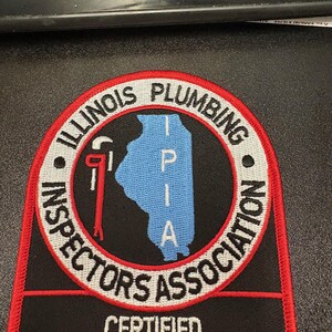 May include: A black and red patch with the text "ILLINOIS PLUMBING INSPECTORS ASSOCIATION" in white. The patch features a blue outline of the state of Illinois and a red wrench. The text "CERTIFIED PLUMBING INSPECTOR" is also displayed.