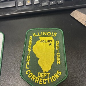 Op de afbeelding: Groen en geel geborduurd patch met de tekst "ILLINOIS CORRECTIONAL DEPT CORRECTIONS" en "JOLIET OFFICER" in een cirkel rond een gele omtrek van de staat Illinois.