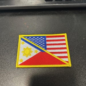 May include: A yellow-bordered patch with the flags of the Philippines and the United States of America sewn together. The Philippine flag is on the left and the American flag is on the right. The flags are sewn together at a diagonal angle.