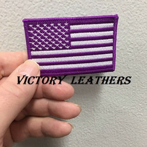 May include: Purple embroidered American flag patch with white stars and stripes. The patch is held in a hand and the text "VICTORY LEATHERS" is visible below the patch.