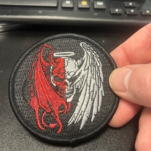May include: A black embroidered patch with a skull in the center. The skull is split in half, with one side showing a red devil's wing and the other side showing a white angel's wing.