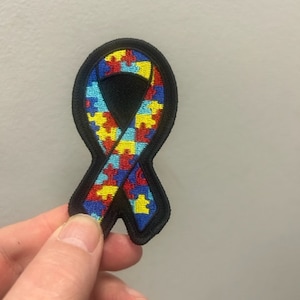 Op de afbeelding: Een zwart omrande, lintvormige patch met een puzzelstukjesontwerp in rood, geel, blauw en groen. De patch wordt door een hand tegen een neutrale achtergrond gehouden. Het ontwerp is een symbool van autismebewustzijn.