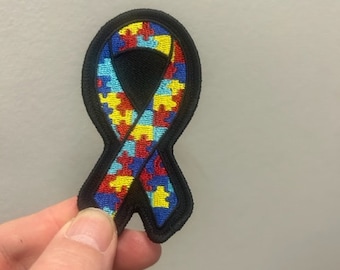 Puede incluir: Un parche con forma de lazo con borde negro y un diseño de piezas de rompecabezas en rojo, amarillo, azul y verde. El parche está sujeto por una mano sobre un fondo neutro. El diseño es un símbolo de concienciación sobre el autismo.
