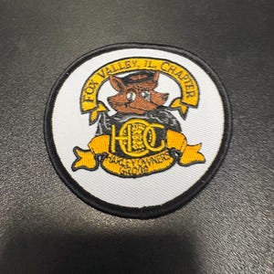 May include: A circular embroidered patch with a white background and a black border. It features a brown fox character wearing a cap, positioned above a yellow banner with the text HOG and HARLEY OWNERS GROUP, and a top banner reading FOX VALLEY, IL, CHAPTER.