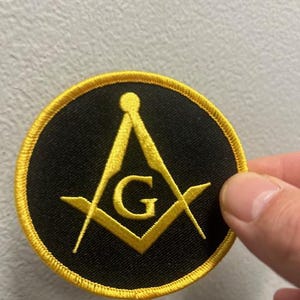 May include: A black and gold embroidered patch featuring a compass and square with a 'G' in the center. The patch is circular and has a gold border.