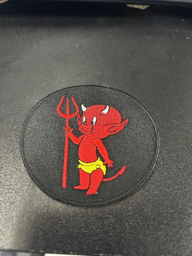 以下が含まれることがあります： 黒い円形のワッペンに、黄色いおむつをした赤い悪魔がフォークを持っている様子が描かれています。