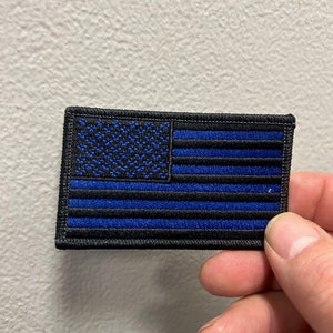 May include: Embroidered patch featuring the American flag with a blue line. The flag has blue stars in the upper left corner and blue stripes. The patch is rectangular with a black border and is held by a hand.