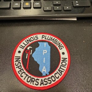 May include: A circular patch with the text "ILLINOIS PLUMBING INSPECTORS ASSOCIATION" in white lettering. The patch features a red border, a black background, and a blue outline of the state of Illinois. A red wrench is also visible.