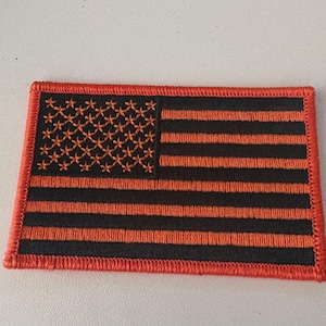 May include: A rectangular embroidered patch featuring a stylized American flag design. The patch is composed of black and bright orange thread, with orange stars in the canton and alternating orange and black horizontal stripes throughout.