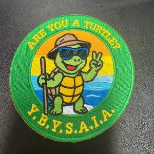 Puede incluir: Un parche bordado circular que presenta una tortuga verde con un sombrero marrón y gafas de sol azules, sosteniendo un bastón y haciendo el signo de la paz. El fondo muestra un sol amarillo y agua azul. El texto dice ARE YOU A TURTLE? Y.B.Y.S.A.I.A.