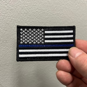 May include: A rectangular patch featuring a stylized American flag design. The flag has a black background, white stripes, and a blue line. The stars are white, and the border is black.