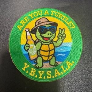 May include: A circular embroidered patch featuring a green turtle wearing a brown hat and blue sunglasses, holding a beer bottle and making a peace sign. The background shows a beach scene with yellow text reading ARE YOU A TURTLE? and Y.B.Y.S.A.I.A.