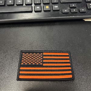 May include: A black and orange embroidered American flag patch. The stars and stripes are orange on a black background.